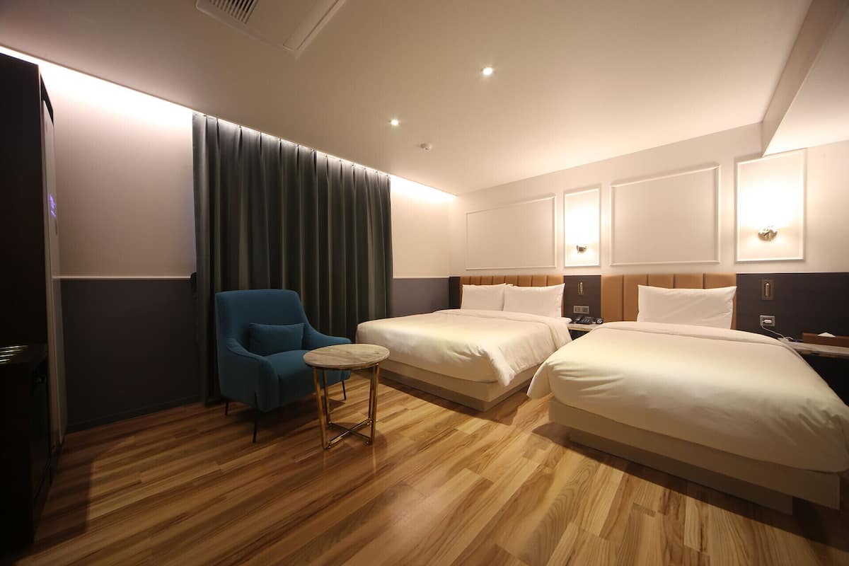 A hotel room with two beds, a small round table, a blue armchair, and a large window with curtains.