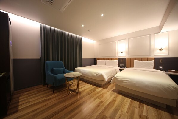 A hotel room with two beds, a small round table, a blue armchair, and a large window with curtains.