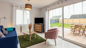 A modern living room with a sofa, a TV, a coffee table, and a pink chair. There is a large sliding glass door leading to an outdoor patio with a table and chairs. - Vacation home Océane by Interhome (Barneville Carteret)