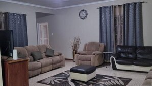 A living room with a beige sofa, a black leather sofa, a coffee table, and a TV on a wooden stand. - 3-Bed House Sleeps 6 with Mountain Views (Ocho Rios)