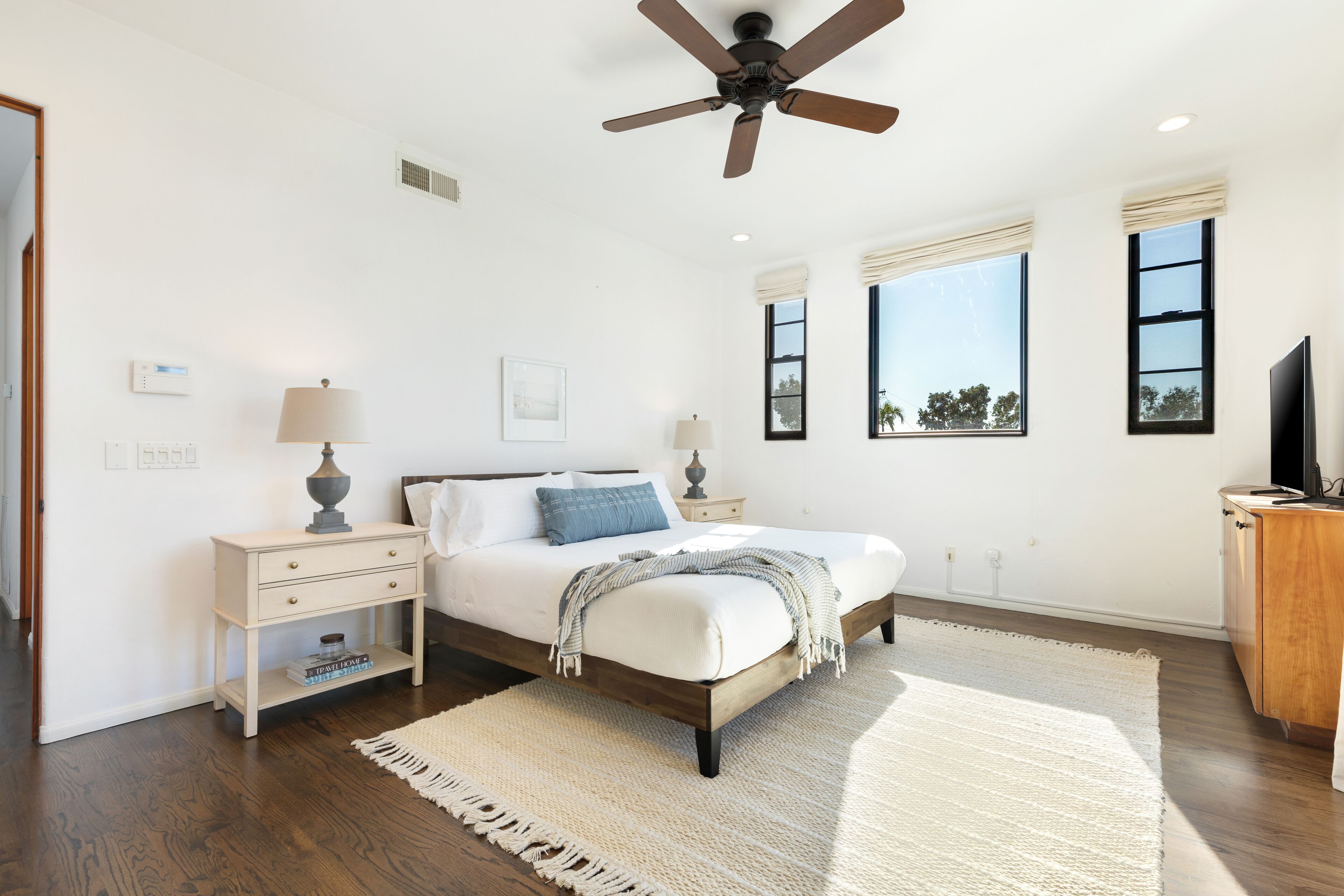 Un dormitorio con cama, mesitas de noche, ventilador de techo y ventanas amplias.