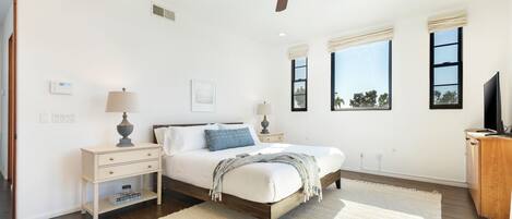 Un dormitorio con cama, mesitas de noche, ventilador de techo y ventanas amplias.