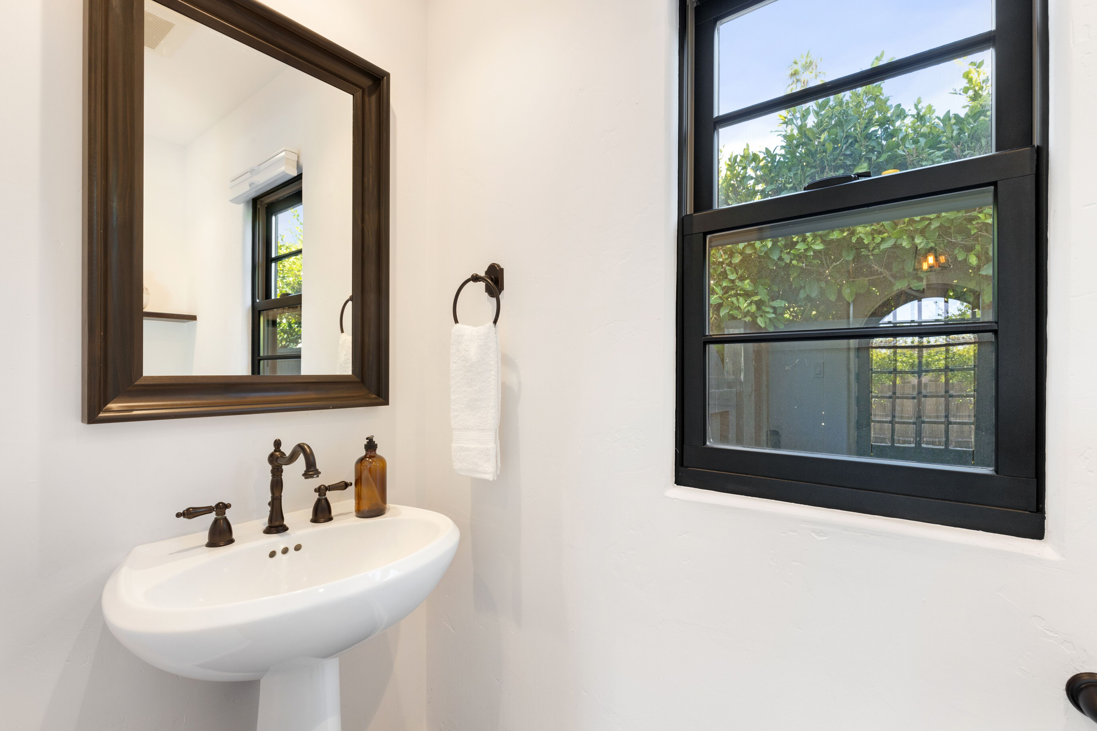 A bathroom with a white sink, a mirror, and a window with a view of greenery outside.