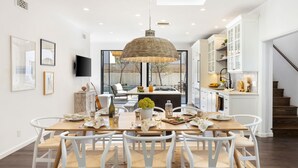 A modern kitchen with white cabinetry, a dining area with a wooden table and white chairs, and a large hanging light fixture.