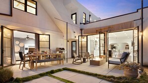 A modern outdoor patio with a dining area, seating, and a kitchen visible through glass doors.