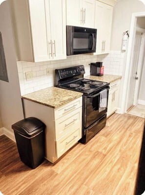 A compact kitchen with white cabinets, a black microwave, a stove, and a trash bin.