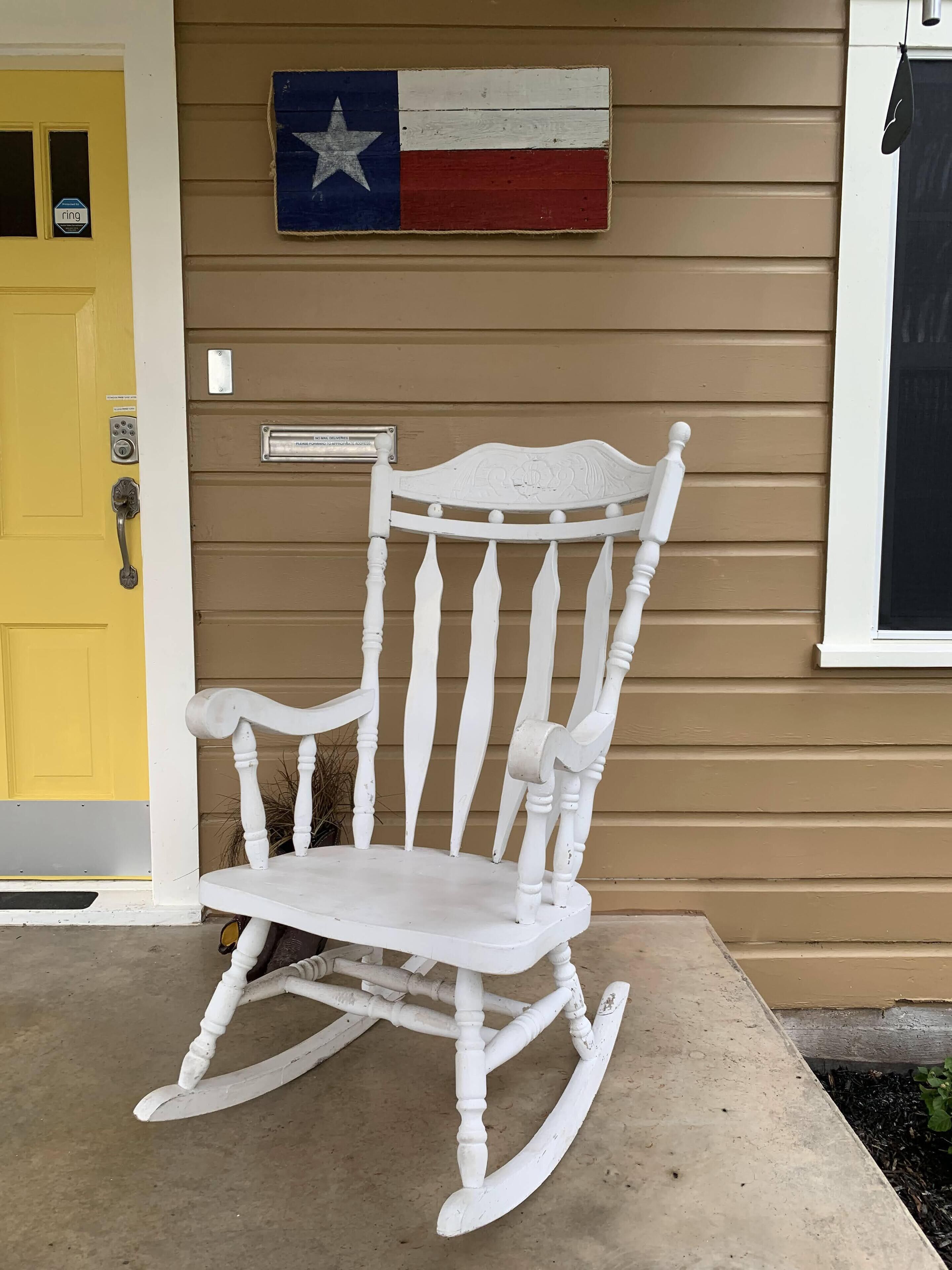 Une chaise berçante blanche est placée devant une maison dont la porte est jaune et sur le mur flotte un drapeau du Texas.
