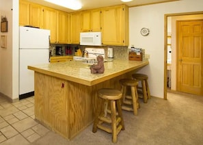 A kitchen with wooden cabinets, a white refrigerator, a microwave, and a granite countertop. There are three wooden stools around the island. - Ski in/ski out across from Austria Hof (Mammoth Lakes)