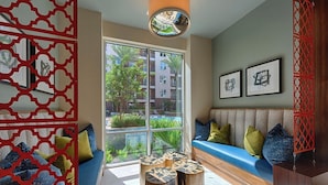 A modern living room with a blue sofa, wooden stools, and a large window offering a view of a pool and buildings. - Downtown Retreat Near Attractions W\ Free Parking (Houston)
