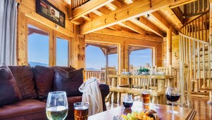 A wooden cabin interior with a sofa, dining table, and chairs. There are glasses of beer and wine on the table. - 100-Mile Views: 3 King Suites I Hot Tub I Game Rm-Pool Table/Arcade/Air Hockey (Sevierville)