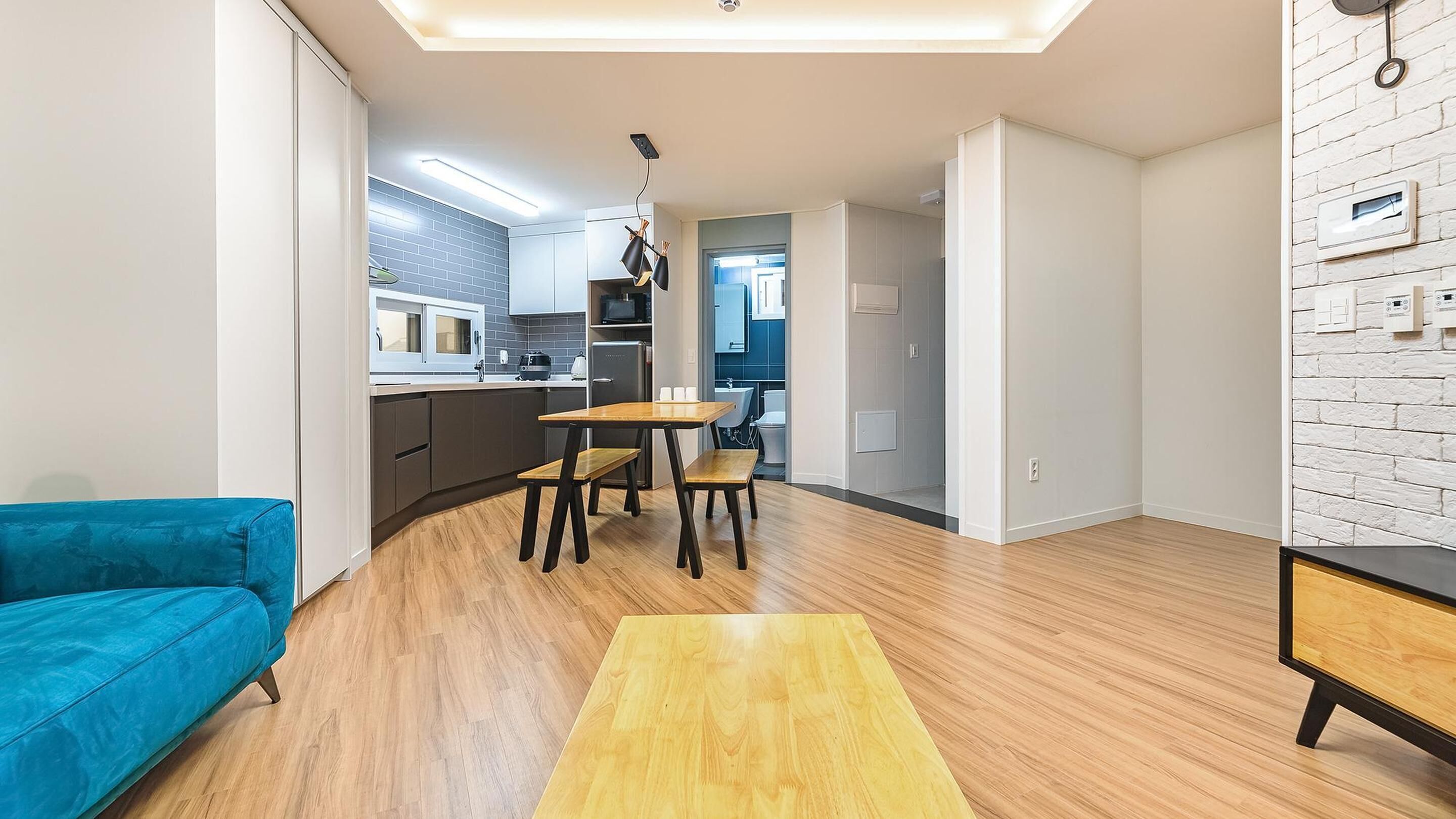 Una sala moderna con un sofá azul, una mesa de comedor de madera y sillas. Se ve una cocina con microondas y armarios al fondo.
