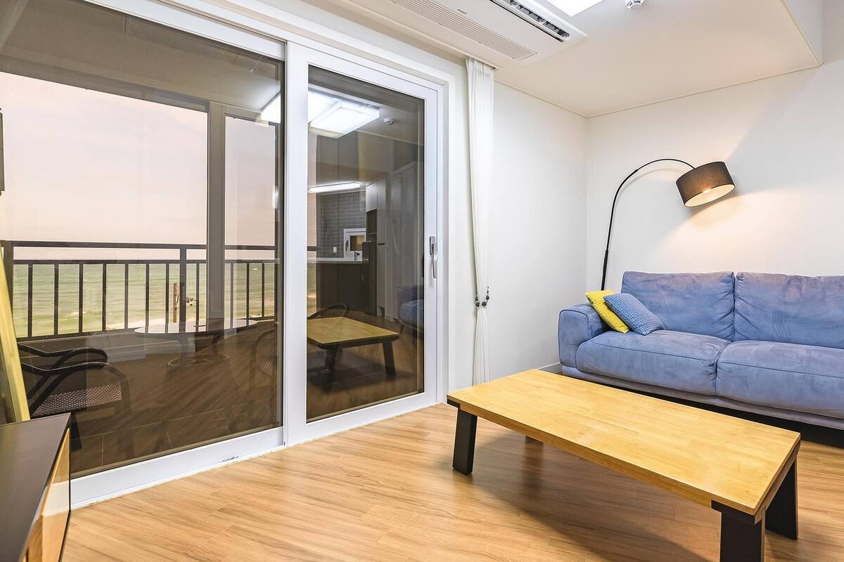 A living room with a sliding glass door leading to a balcony, a blue sofa, a wooden coffee table, and a floor lamp.