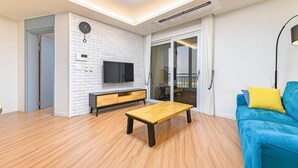 Una sala de estar moderna con una mesa de centro de madera, un sofá azul con cojines amarillos, un televisor de pantalla plana montado en una pared de ladrillo blanco y un puertas correderas de vidrio que dan a un balcón.