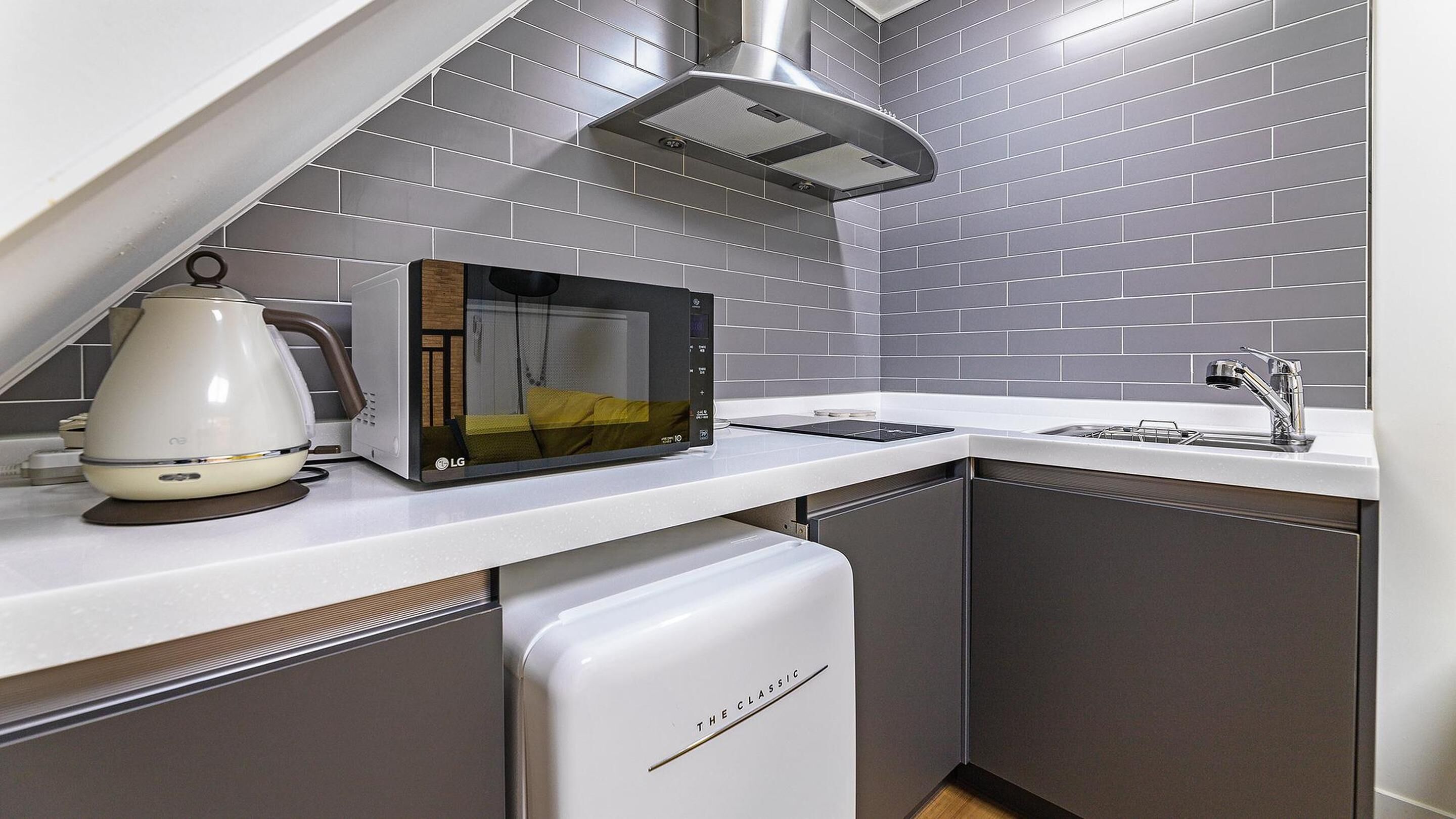 A compact kitchen with grey tiled backsplash, a built-in microwave, a kettle, and a sink with a modern faucet.