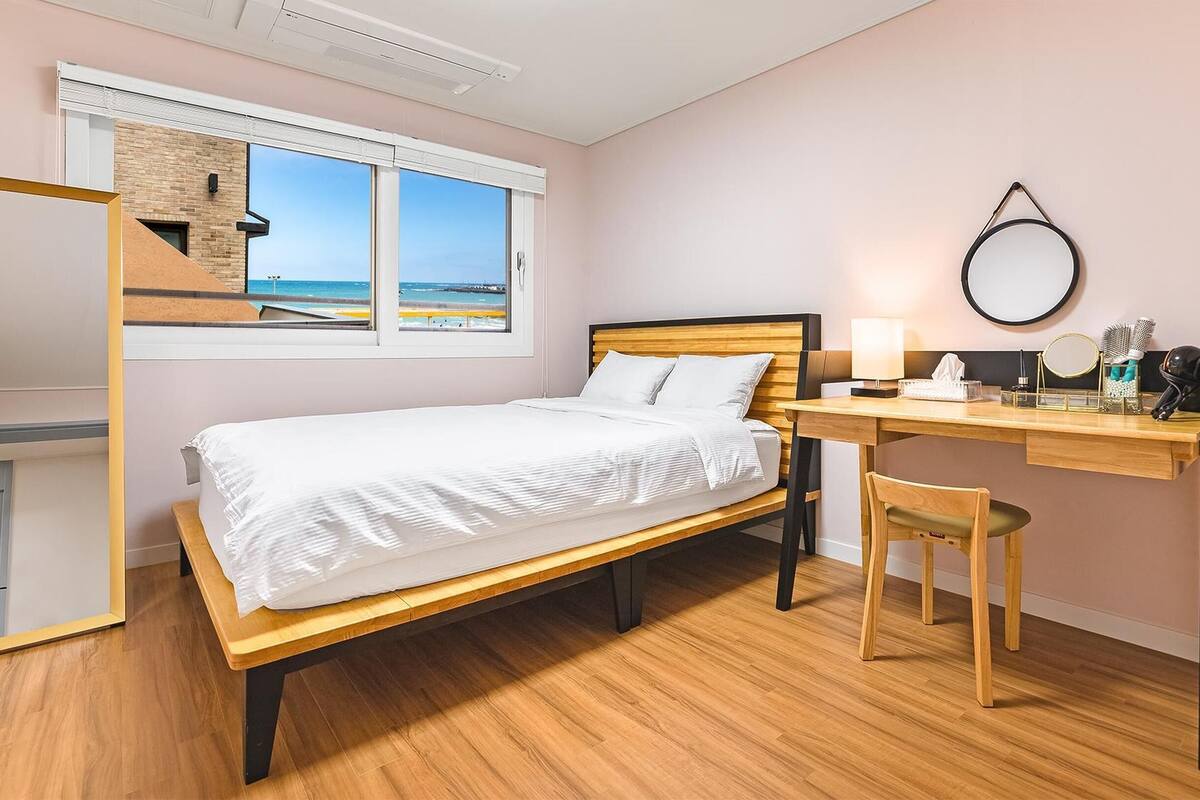 A bedroom with a bed, a desk, and a chair. There is a window with a view of the beach.