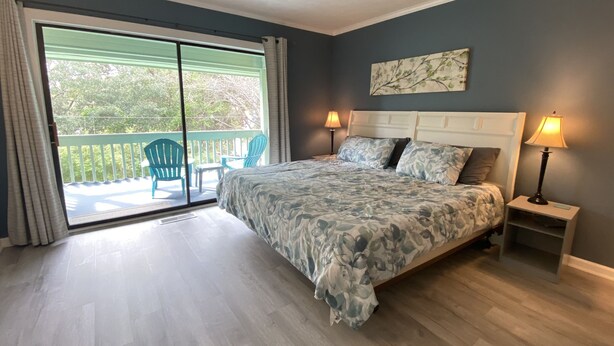 Een slaapkamer met een groot bed, twee bedlampen, uitzicht op een balkon met stoelen, en een schilderij aan de muur.