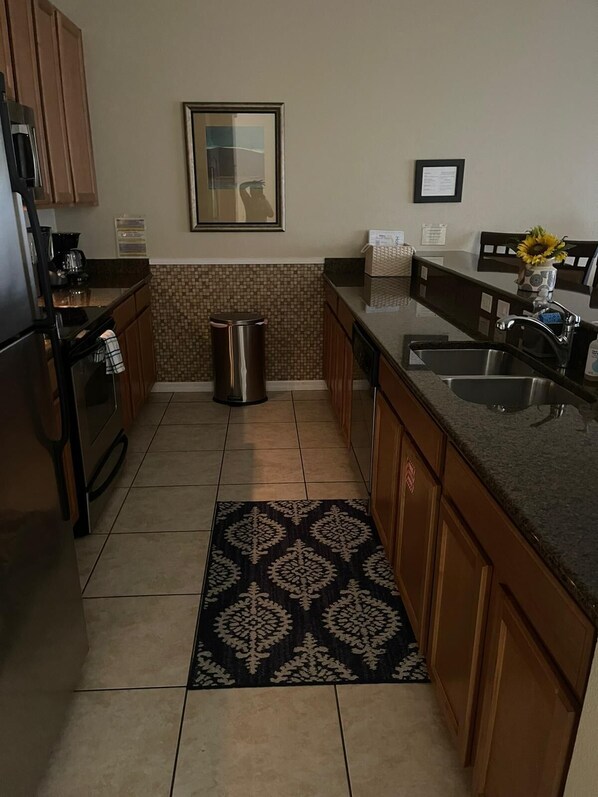 Una cocina con armarios de madera, un bote de basura de acero inoxidable, salpicadero de azulejos y un tapete estampado.