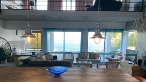 A modern living room with a wooden dining table, sofas, and hanging chairs. Large windows provide a view of the outdoors. - Pet-Friendly Villa Azores SunriseHouse with Sea View, Pool, Jacuzzi, Wi-Fi, Terrace & Garden (Pedreira)