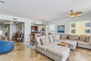 A spacious living room with a large beige sectional sofa, a blue armchair, and a dining area with wooden chairs and a table.