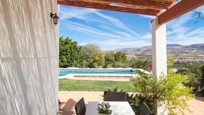 Une terrasse couverte avec une table et des chaises, une piscine en arrière-plan et une vue sur le paysage.