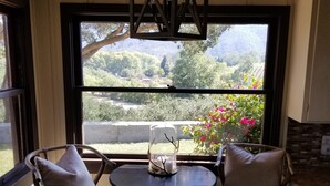 Ein kleiner Essbereich mit einem runden Tisch und zwei Stühlen, einer Hängeleuchte und einem Fenster mit Blick auf Grünflächen und Hügel.