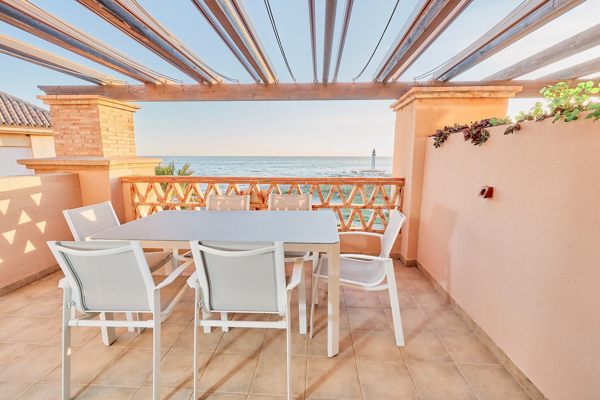 A covered outdoor patio with a table and chairs, a view of the sea, and a lighthouse in the distance.