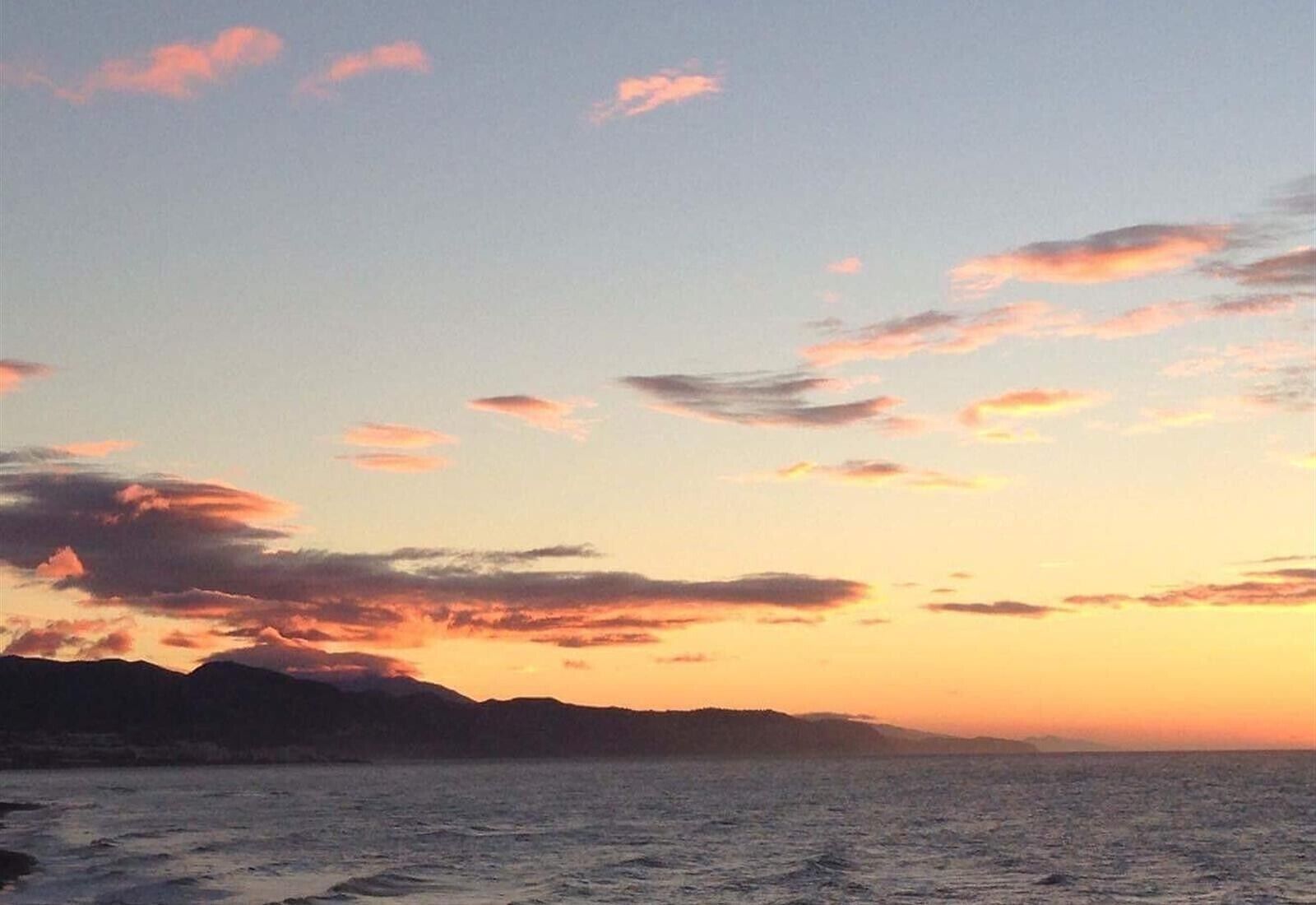 Atardecer costero con nubes, mar tranquilo y tierra a lo lejos.