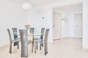 Een moderne eethoek met een glazen tafel en metalen stoelen, een vaas met bloemen en een witte deur op de achtergrond.