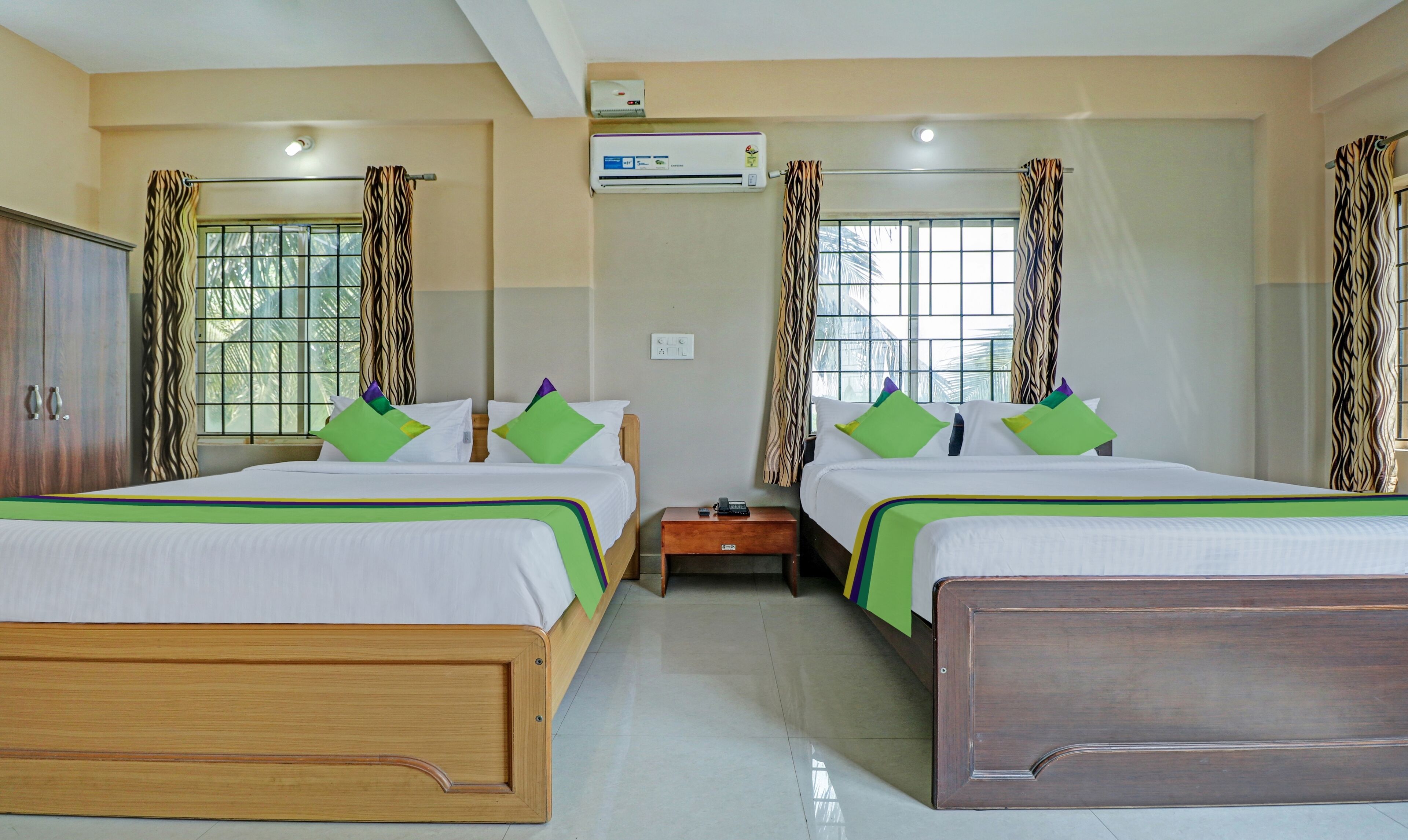 A hotel room with two beds, each with green and purple pillows, a wooden headboard, and a small wooden nightstand with a phone and remote control.