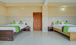 Habitación de hotel con dos camas, cada una con cabecero de madera, ropa de cama blanca y almohadas verdes y moradas. Hay mesitas de noche con lámparas y un banco de madera al pie de las camas.