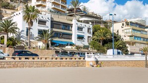 A coastal building with balconies, a stone wall, parked cars, and palm trees.