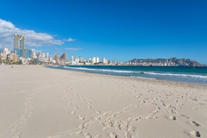 A beach with footprints leading to the water, a city skyline in the background, and mountains in the distance.