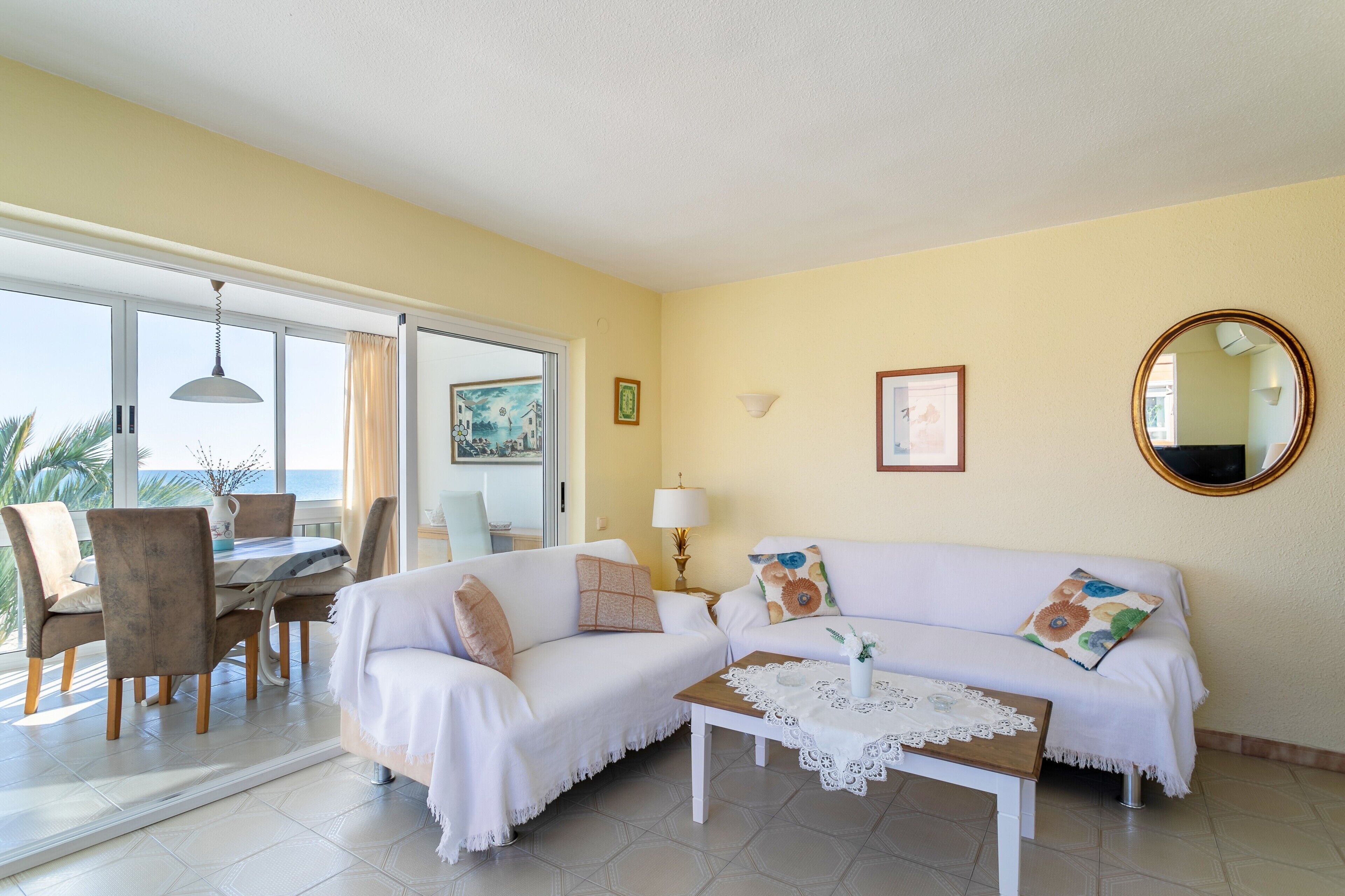 Una habitación con un sofá blanco, una mesa de comedor con sillas, una mesa auxiliar y vistas a través de un ventanal.
