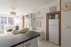 Een moderne keuken met een zwart aanrecht, witte kasten, een magnetron en een koelkast. Er is een eethoek met een tafel en stoelen, en een raam met uitzicht op gebouwen.
