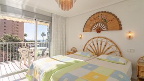 Une chambre d’hôtel avec un balcon, un lit doté d’une tête de lit décorative, un ventilateur mural et une vue sur des palmiers et des immeubles.