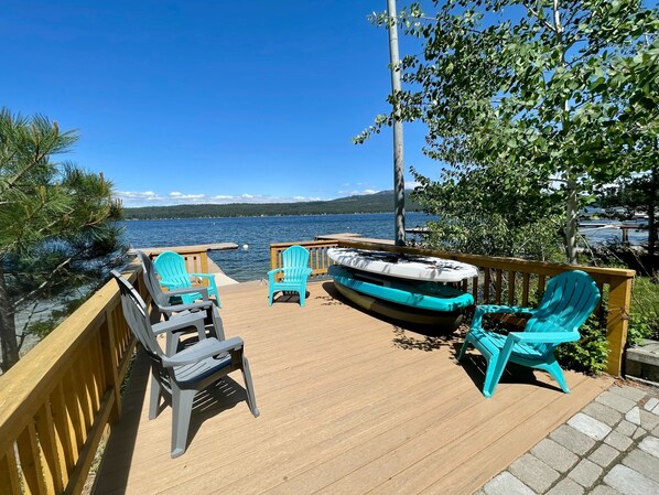A wooden deck with outdoor seating, including chairs and a boat, overlooking a body of water with a distant shoreline.