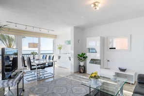 A modern living room with a glass coffee table, a sofa, a TV, and a dining area with chairs and a table.