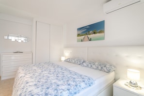 A bedroom with a bed, a nightstand, a wall-mounted air conditioner, a bedside lamp, and a painting of a beach scene. - Apartment Casa Esmeralda 2 on the Beach with Balcony, A/C & Wi-Fi (Benidorm)