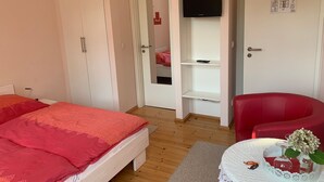 Une chambre à coucher avec un lit, un fauteuil rouge, un téléviseur fixé au mur et une porte donnant sur une autre pièce.