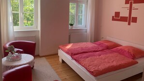 Une chambre à coucher comprenant un lit, une chaise, une petite table ornée d’un objet décoratif et deux fenêtres.