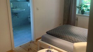 A bedroom with a bed, a bedside table with a vase and remote control, a window with a plant, and a bathroom visible through an open door. - Single room 4 - Loensches Gut (Görlitz)
