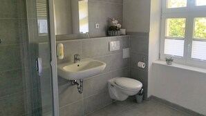 A modern bathroom with a glass shower enclosure, a sink with a mirror, a toilet, and a window with blinds. - Single room 4 - Loensches Gut (Görlitz)