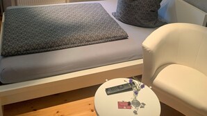 A bed with a patterned blanket and pillows, a bedside table with a remote control and a card, and a white chair. - Single room 4 - Loensches Gut (Görlitz)