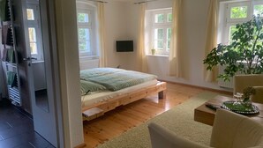 Ein Schlafzimmer mit einem hölzernen Bett, einem an der Wand montierten Fernseher, einem Blumentopf und einem Fenster mit Vorhängen.