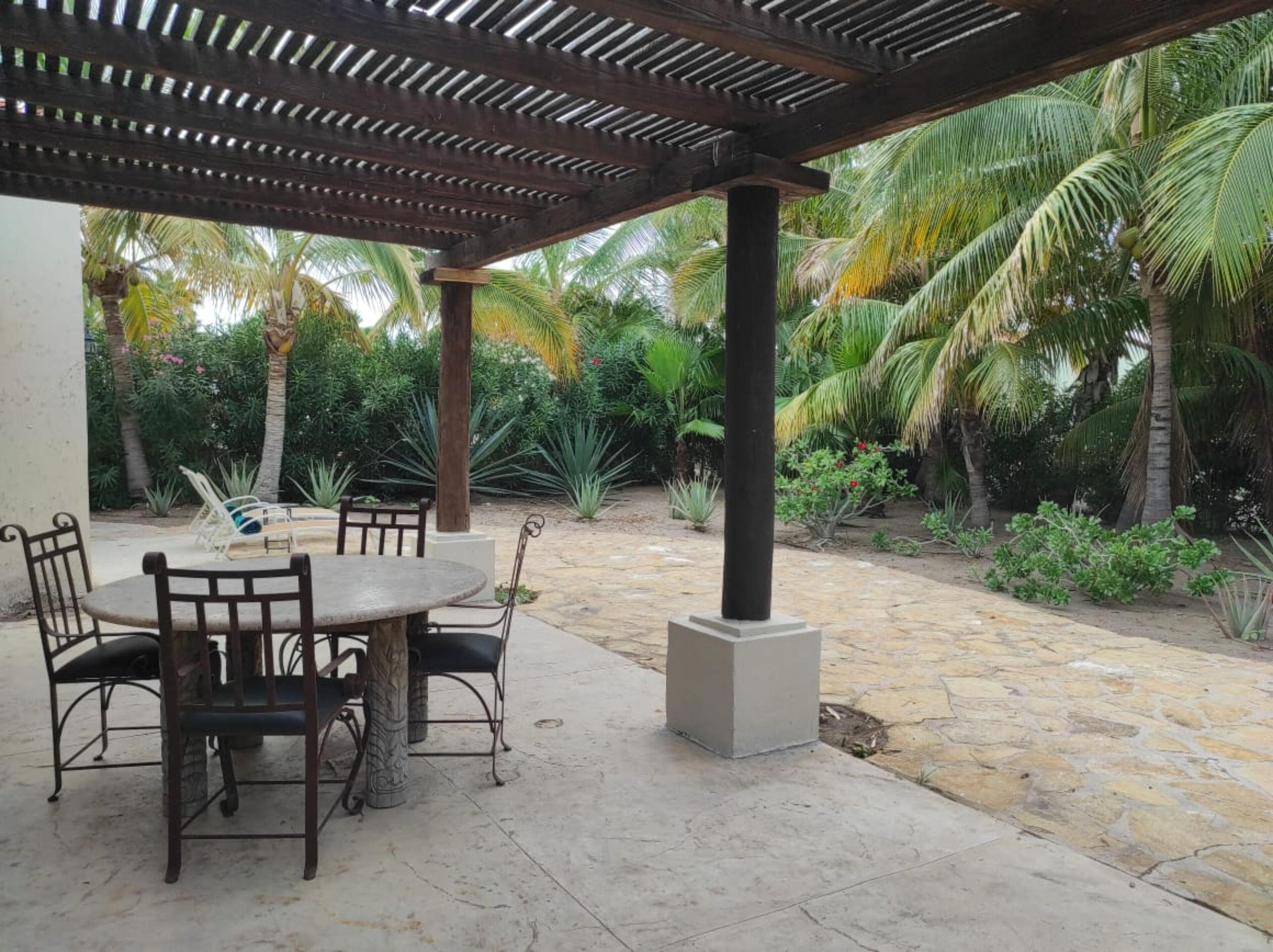 An outdoor patio with a round table and chairs, surrounded by palm trees and a pergola.