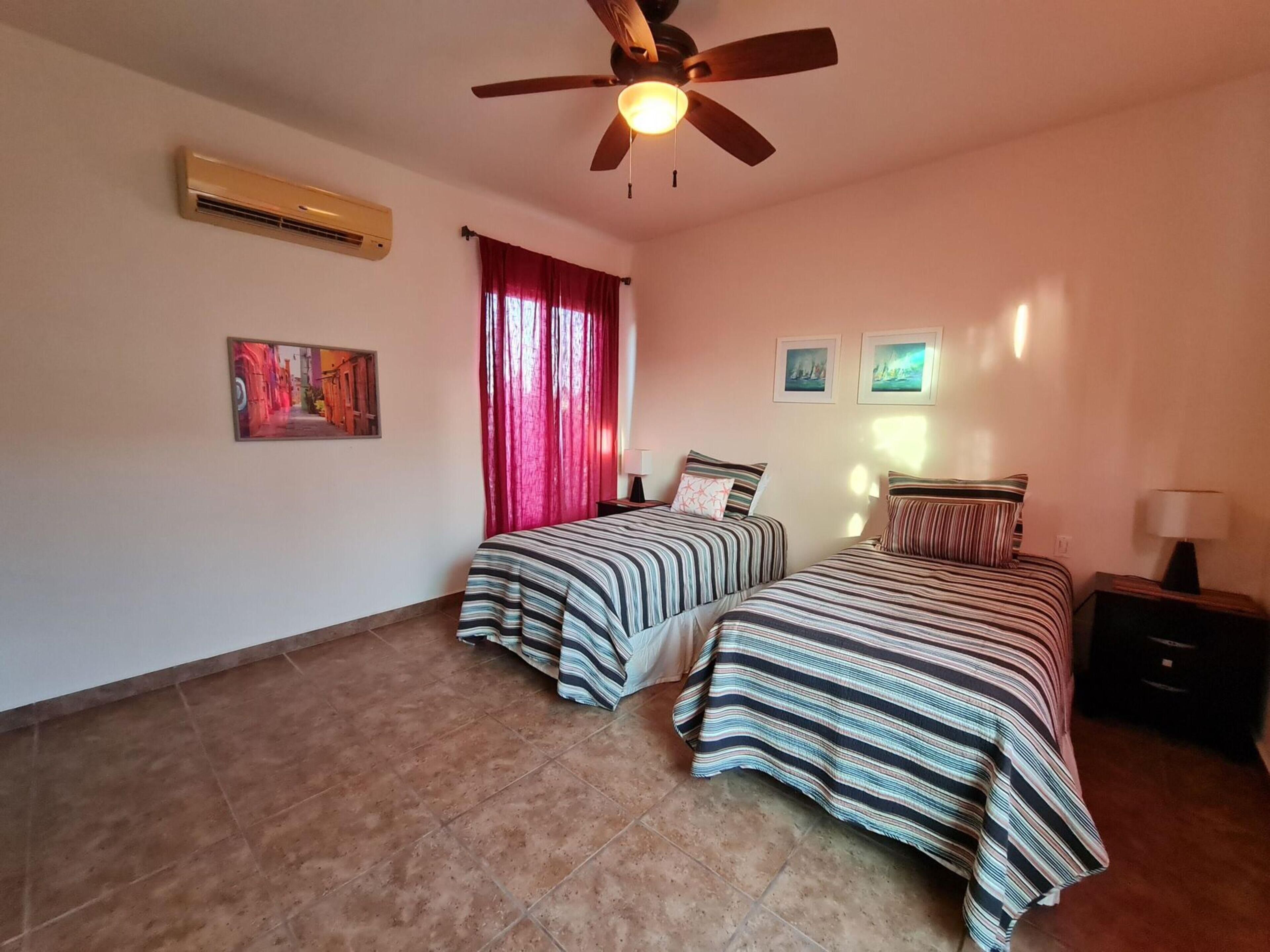 Ein Schlafzimmer mit zwei Betten, einem Deckenventilator, einer Klimaanlage und zwei gerahmten Bildern an der Wand.