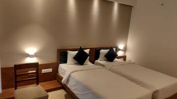 A hotel room with two single beds, a wooden nightstand with a lamp, a wooden chair, and a small table.