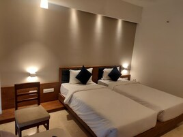 A hotel room with two single beds, a wooden nightstand with a lamp, a wooden chair, and a small table.