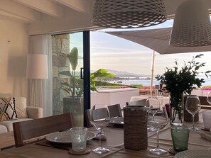 Un coin repas avec une table dressée pour deux personnes, une vue sur la mer et un mur en pierre.