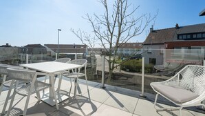 Un patio al aire libre con una mesa y sillas, barandilla de vidrio y vista a edificios residenciales.